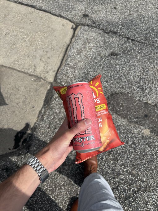 A hand holds a bag of Monster Energy "Pipeline Punch" chips with 10g of protein over a sunlit sidewalk, capturing the 24-hour energy rush behind the bullish NFT enthusiasm.