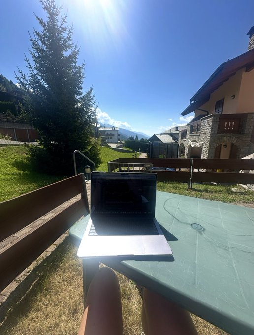 A laptop sits open on a green outdoor table in a sunny mountain village setting, with a person's legs visible in the foreground and stone buildings nearby.