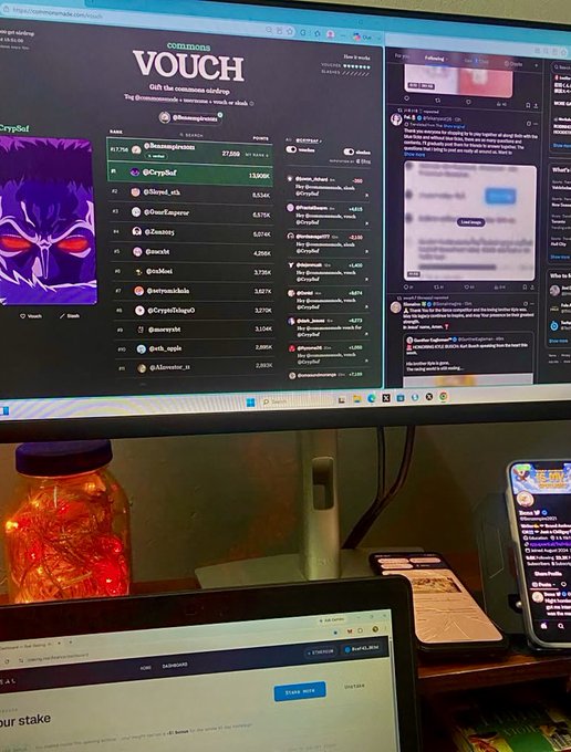 A computer monitor displays the VOUCH platform leaderboard showing various usernames, numbers, and NFT avatars, with a jar of orange lights and a smartphone visible on the desk below.