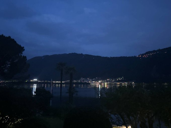 A serene twilight view of Lugano's lake and mountains, with city lights reflecting on the calm water under a deep blue sky, capturing the quiet beauty of dawn breaking over the city. The image aligns with the post's sentiment, showcasing the tranquil moment when Lugano awakens, its lights gradually giving way to the new day.