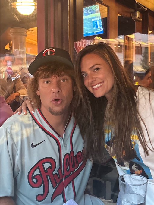 Comedian Theo Von, wearing a "TC" cap and a Phillies baseball jersey, poses with a smiling brunette woman inside a restaurant with a TV visible in the background. The image accompanies a post in which a woman (likely the person pictured) suggests podcast guest ideas to Theo while he waits for Bitcoin's price to recover.