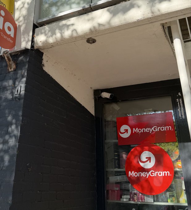 The exterior entrance of a MoneyGram location displays two prominent red MoneyGram signs on the glass door and window, with a small security camera mounted above. The image aligns with the post by illustrating the physical storefront where one can convert euros to Stellar (XLM) using MoneyGram's cash-to-crypto service, bypassing traditional banking channels.