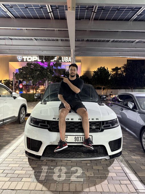 A young man in a black t-shirt and shorts sits on the hood of a white Range Rover, pointing at the camera, parked under a covered area with a 'Top Golf' sign visible in the background.