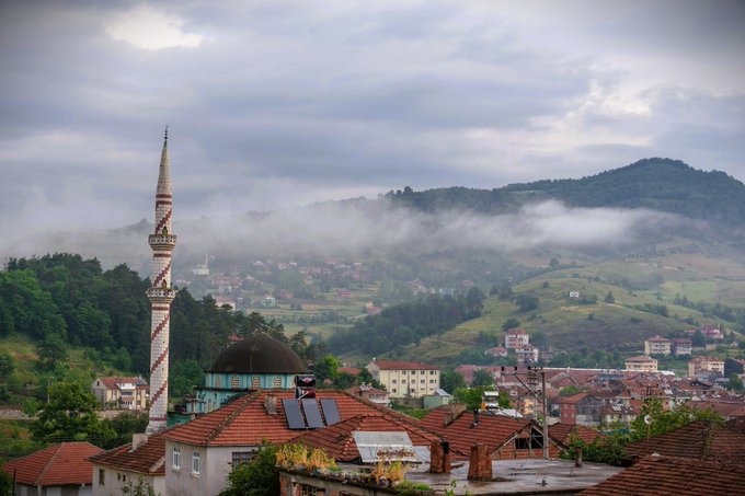 A misty mountain town features a prominent mosque with a tall, spirally-striped minaret and domed roof, surrounded by red-tiled rooftops and nestled among green hills.