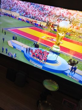A TV screen displays Spain's World Cup trophy celebration ceremony on the pitch, with players gathered around the golden FIFA World Cup trophy, matching the post's congratulations to Spain as world champions.
