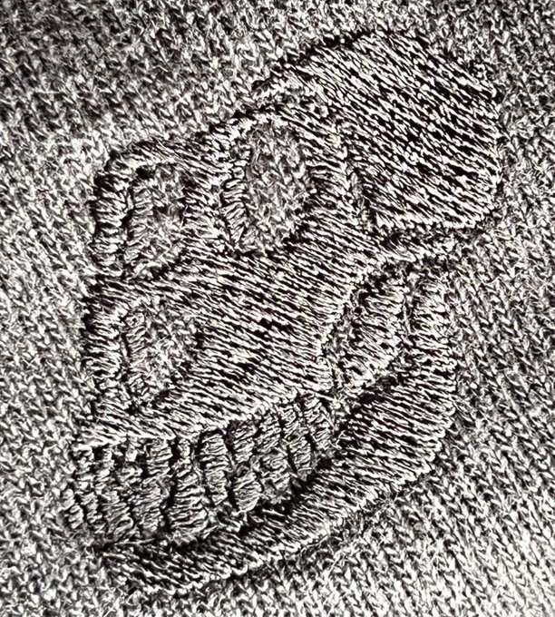 A textured gray surface resembling a woven fabric or rough material, featuring a large raised design that appears to form letters or numbers in the center.