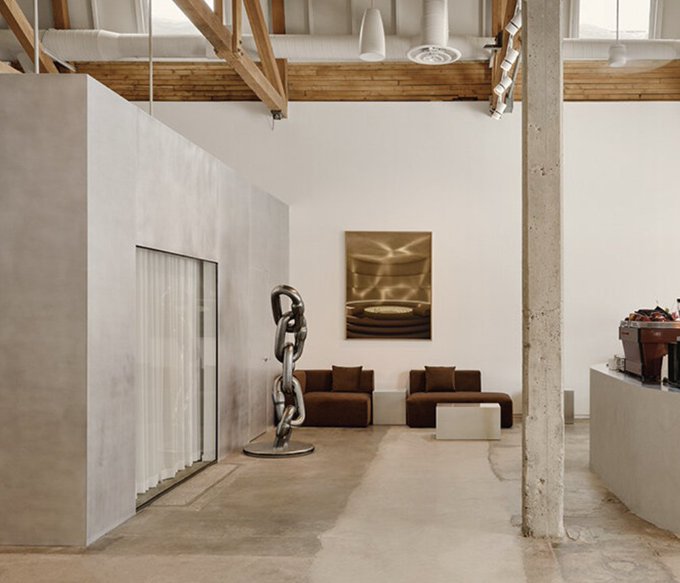 A spacious loft-style interior features exposed wooden beams, concrete columns, polished cement floors, and a striking metallic sculpture beside two brown armchairs. The post playfully jokes that surrounding oneself with this elegant, art-filled space is a form of "choosing who to surround myself with" — suggesting the room itself is the chosen companion.
