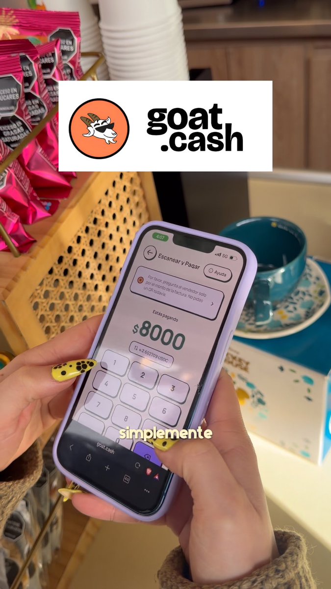 A hand with yellow polka-dot nails holding a phone displaying a crypto payment app screen showing '$8000' with a 'goat.cash' logo overlay, set against a background of snacks and a teal bowl.