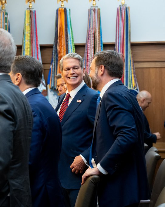 A smiling man in a dark blue suit with a red striped tie and American flag pin laughs warmly while conversing with other suited attendees in a formal government chamber adorned with ceremonial flags. The image contrasts with the post's message about a cryptocurrency fund claiming to donate $50k to children's accounts, as the professional, official setting suggests traditional political or institutional activity rather than the tokenized charity initiative being promoted.