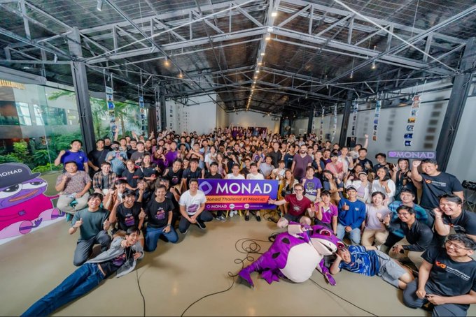 A large group photo of the Monad community event, with attendees posing around a purple 'MONAD' banner and a purple mascot character lying in the foreground.