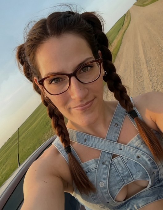 A selfie of a woman with brown braided hair wearing glasses and a denim top, taken outdoors with a dirt road and green field visible in the background.