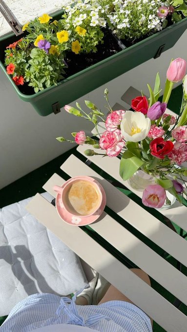 An overhead lifestyle photo of a balcony or patio scene featuring a pink cup of coffee, a vase of fresh flowers including tulips and carnations, and a window box planter with yellow and purple pansies.