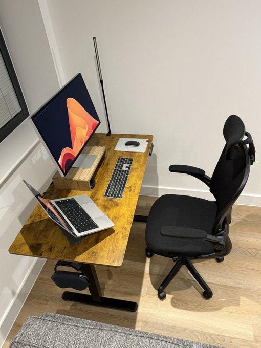 A minimalist home office setup featuring a wooden desk with a desktop monitor displaying a colorful wallpaper, a laptop, keyboard, and a black ergonomic office chair.