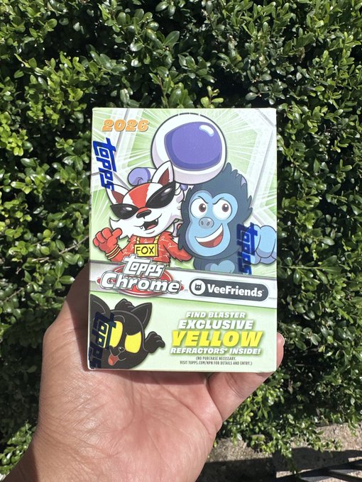 A hand holding a sealed 2026 Topps Chrome trading card pack featuring VeeFriends characters, advertising "BLASTER EXCLUSIVE YELLOW REFRACTORS INSIDE", photographed against a green hedge.