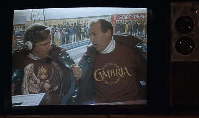 A scene displayed on an old television showing two men wearing maroon sweatshirts; one wears a shirt with an angel graphic, and the other wears a 'CAMBRIA' shirt and holds a microphone.