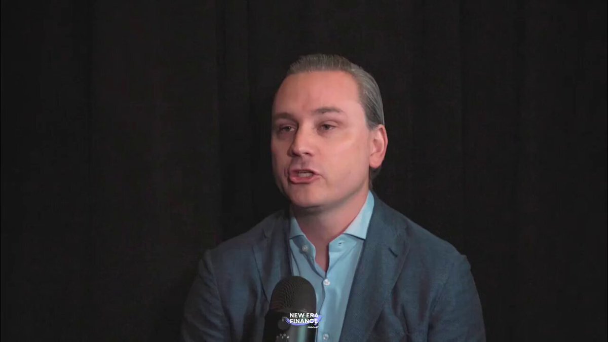 The image shows a man sitting behind a microphone, giving an interview or speaking into it. He appears to be dressed professionally with a suit jacket and tie. The background of the image is black, which could suggest that he is indoors. There are no other people in the scene besides the man speaking through the microphone.