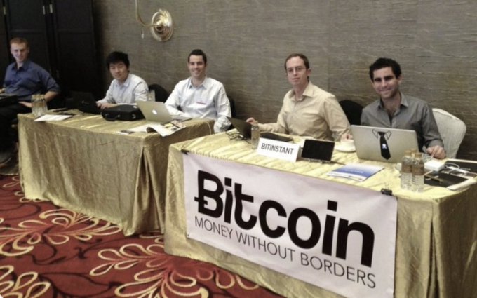 Five men sit at a long table with laptops during a Bitcoin conference, behind a banner reading 'Bitcoin MONEY WITHOUT BORDERS' with a 'BITINSTANT' nameplate visible.