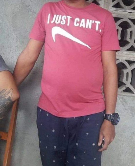 A person wearing a pink Nike t-shirt with the text "I JUST CAN'T." and the Nike swoosh logo, paired with dark patterned shorts and a wristwatch, stands next to another person with a visible tattoo.