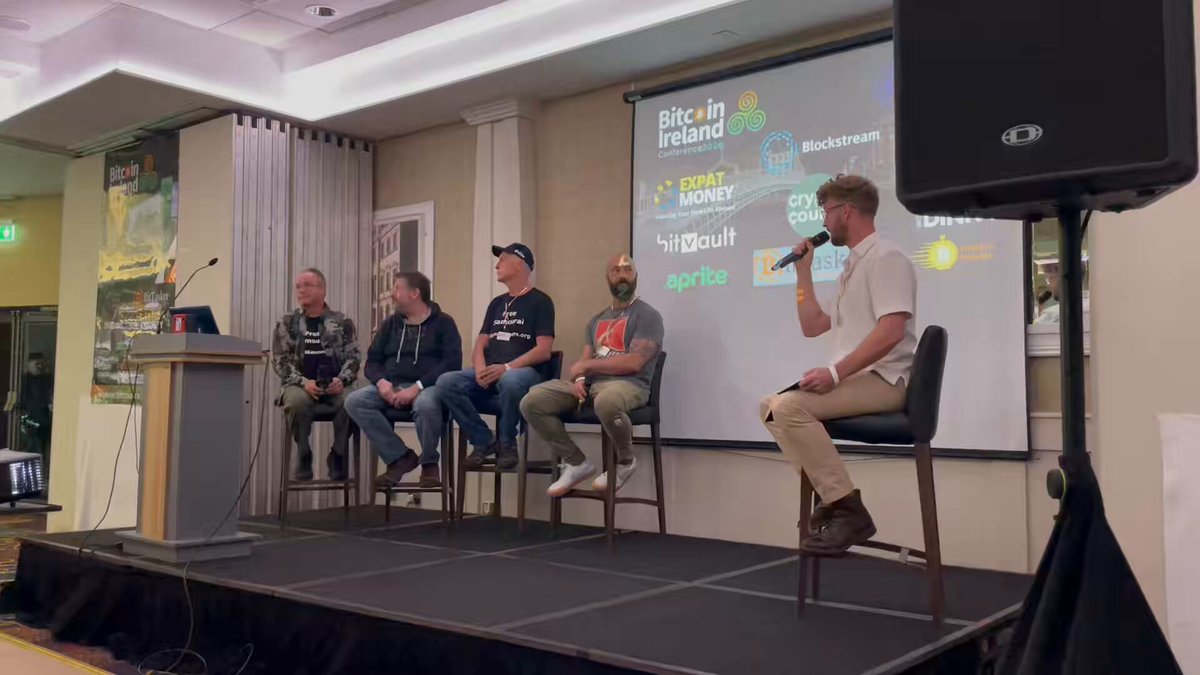 A man stands behind a podium giving a speech at an event with four other men sitting in chairs in front of him. The room has a stage set up for presentations or discussions. In the background, there are posters hanging on the wall that read "We must stop the persecution of Bitcoin and privacy developers from witch hunts".