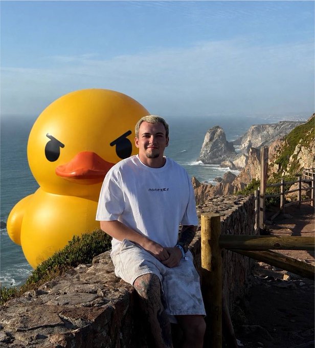 A young man poses with a giant yellow rubber duck by a scenic coastal cliff, announcing his role as an ambassador for Duel Duck (@duel_duck), with his white shirt displaying the brand name "norcees." The playful duck visual ties into the platform's theme of fun prediction battles alongside @denko0x.