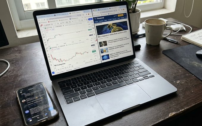 A workspace setup featuring a MacBook displaying TradingLite charts and Financial news, with a smartphone showing social media, a coffee mug, and a notebook on a wooden desk.