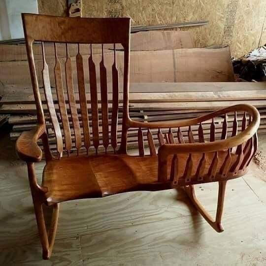 A handcrafted wooden chair with a slatted, curved backrest and armrests sits in a workshop with wood scraps in the background.