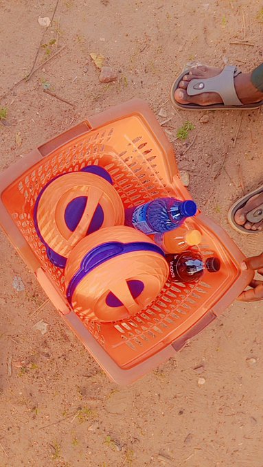 A top-down view of an orange plastic basket on sandy ground containing two round bread-like items with purple centers, a blue water bottle, and a brown soda bottle, next to a person's sandaled foot.