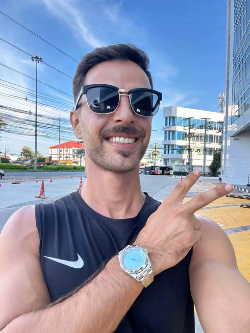A smiling man wearing sunglasses and a black Nike tank top poses outdoors in a parking lot, gesturing toward a silver wristwatch on his wrist.