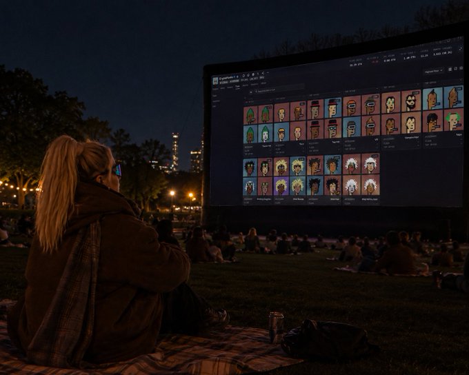 A woman with blonde hair in a ponytail and a brown jacket sits on a blanket at an outdoor nighttime movie screening, watching a large screen displaying an NFT marketplace with various cartoon character avatars.