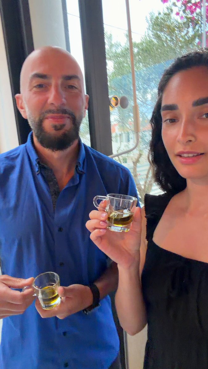 In the image, a man wearing a blue shirt holds a glass of wine while standing next to a woman who is also holding a glass of wine. The two people are positioned close together, with both of them looking at the camera. The background features a window and a door, suggesting that they might be in an indoor setting like a living room or dining area.