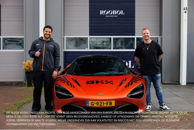 Two men stand on either side of an orange sports car (license plate G-921-FB) in front of a 'ROOBOL EXCLUSIVE CARS' dealership, with Dutch text at the bottom disclosing that OKX is a marketing name for OKX Europe Ltd.