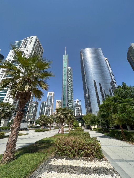 A bright cityscape featuring modern skyscrapers and a tall green glass tower, viewed from a landscaped plaza with palm trees under a clear blue sky.