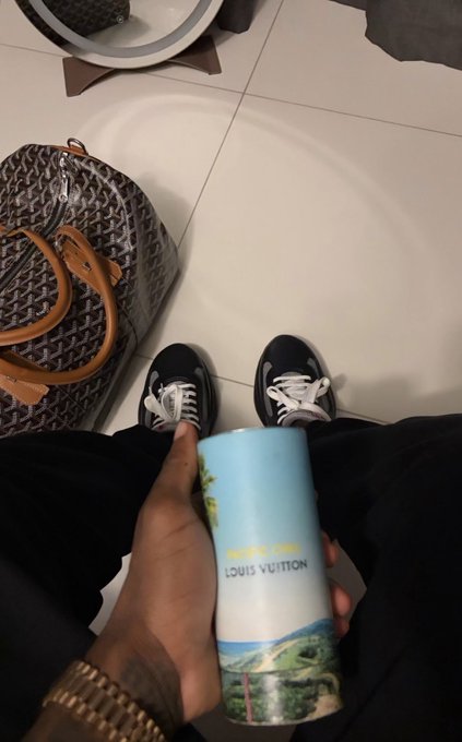 A first-person view looking down at sneakers, a designer duffle bag, and a tumbler cup labeled 'LOUIS VUITTON' being held in hand.