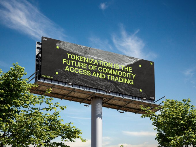 A billboard from metals.io and GM (presumably the brand it advertises for) is mounted on a pole against a partly cloudy blue sky, displaying the text "TOKENIZATION IS THE FUTURE OF COMMODITY ACCESS AND TRADING" in bold yellow letters. The image directly visualizes the post's message, showcasing a large-scale advertisement promoting the concept of tokenization in commodity markets.