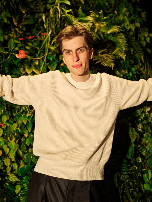 A young man with tousled brown hair poses with arms outstretched against a lush green foliage backdrop, wearing a cream-colored knit sweater. The caption juxtaposes the carefree, confident pose with the boastful claim about getting rich, suggesting an aspirational or satirical tone about wealth and success.