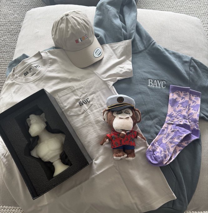 The image features a variety of merchandise, including clothing items such as t-shirts and hats, along with a stuffed monkey toy. The monkey appears to be wearing a hat that says "BAYC", indicating its connection to the BAYC brand. The clothing items are displayed on a bed or couch, creating an inviting atmosphere for potential buyers. The post about the merchandise includes text such as "Thanks @mfigge" and "Now I can rep the BAYC in every video Opened it live at 40:40".