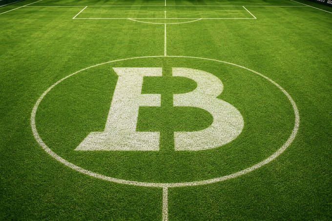 The image features a soccer field with a white logo of the letter "B" drawn on the green grass, surrounded by white lines. The text "BM" is also present within the logo. The background of the image shows a soccer pitch or field, indicating that this could be an advertisement for Bitcoin or a related cryptocurrency event.