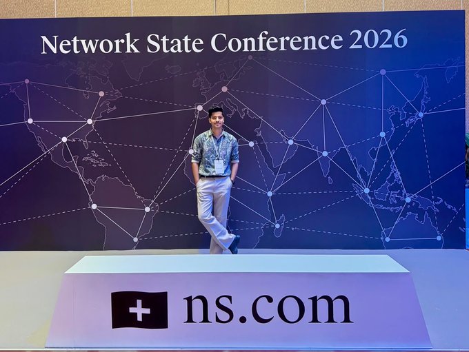 A young man posing at the Network State Conference 2026 in front of a world map backdrop with network connection lines and a large ns.com sign in the foreground.