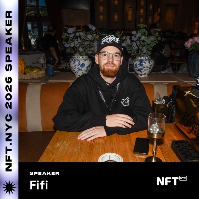 The image features a man wearing glasses sitting at a wooden table with a glass of wine in front of him. The background includes potted plants and vases, suggesting that they are in an indoor setting like a restaurant or bar. There is also text on the image that reads "Speaker @NFT_NYC" and "@doginaldogs for leading the way." This indicates that the man sitting at the table might be a speaker or a guest of honor at an event, possibly related to dogs or pet-related topics.