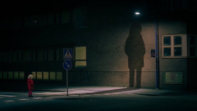 A cinematic nighttime scene showing a small child in a red jacket standing at a crosswalk, gazing up at a giant shadowy figure cast against a building wall under a streetlamp.