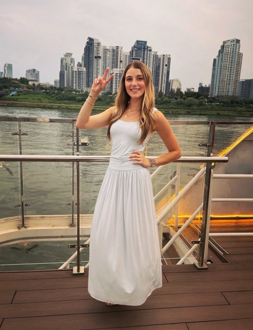 A young woman in a flowing white dress poses with a peace sign on a riverside deck, with Seoul's high-rise skyline visible across the water—greeting her followers with "hello from seoul."