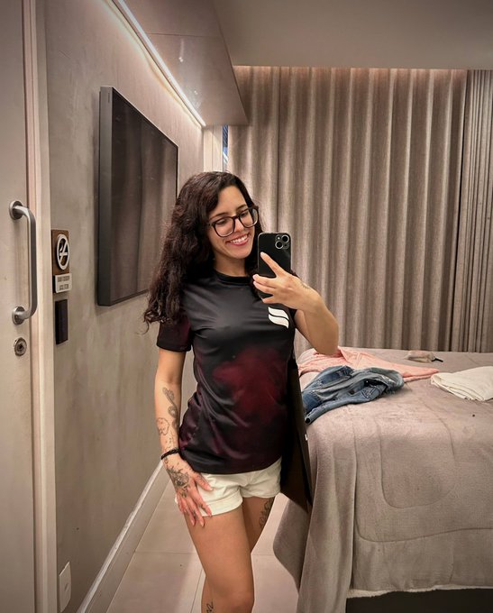 A woman with curly hair and glasses smiles while snapping a mirror selfie in a modern bedroom, holding her phone up to capture the shot. Her relaxed, casual outfit and bright morning energy fit the post's cheerful "Good morning, let's mint today!" vibe.