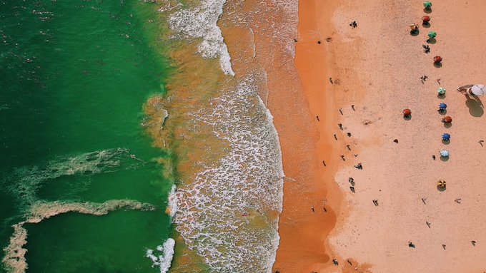 An aerial view of a beach with green ocean water meeting orange sand, where beachgoers with colorful umbrellas dot the shoreline.