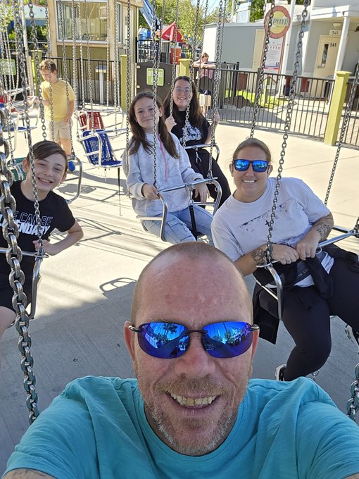 A selfie of a bald man wearing blue reflective sunglasses and a turquoise shirt, with his family smiling on swings at what appears to be an amusement park.