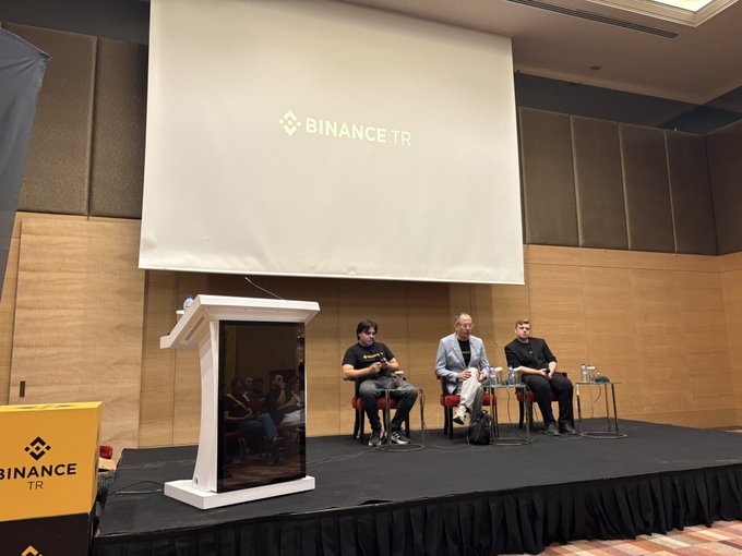 Three people sit on stage at a 'BINANCE TR' event with the logo projected on a screen behind them, with one man speaking at a podium labeled 'BINANCE TR'.
