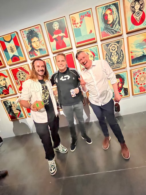 Three smiling men posing together in an art gallery with colorful pop-art style framed portraits covering the wall behind them, one holding a can and another holding a bottle.