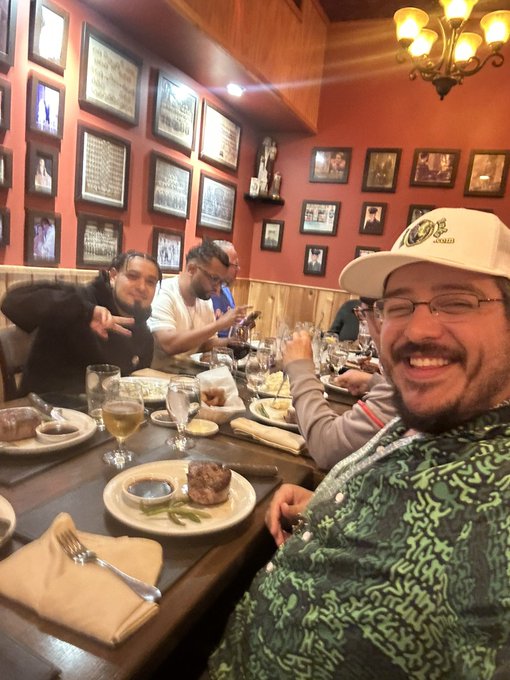Two men posing for a selfie at a restaurant table with food and drinks, the man in front wearing a green patterned shirt and a white 'ACADEMY' cap.