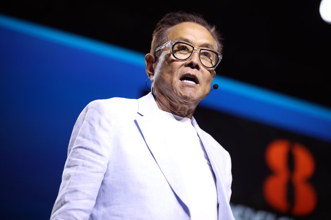 The image features an older man wearing glasses and a white suit standing on stage with his mouth open as if he's speaking or giving a presentation. The background of the image shows a blue wall with a red logo, which appears to be related to the post about Robert Kiyosaki discussing the potential value of gold, silver, Bitcoin, and Ethereum.