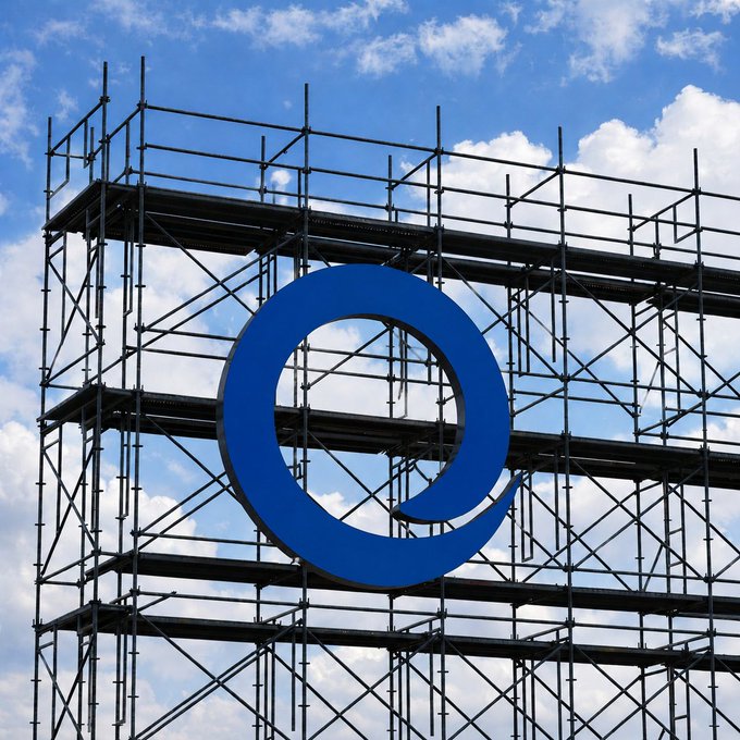 A large blue letter 'Q' displayed on metal scaffolding against a partly cloudy sky.