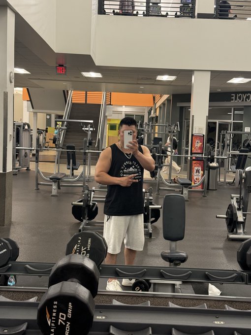 A mirror selfie taken in a gym, showing a man in a black tank top and white shorts holding a phone, with a dumbbell rack and various workout equipment visible in the background.